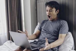 förvånad och chockad asiatisk man arbetar med sin bärbara dator på sin mysiga säng. begreppet framgångsrik frilansar livsstil. foto
