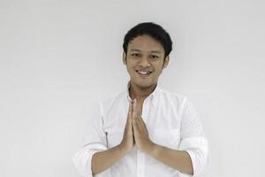 ung asiatisk man klädd i vit skjorta ger hälsning händer på med ett stort leende på läpparna. indonesisk man på grå bakgrund. eid mubarak foto