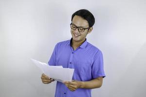 ung asiatisk man är leende och glad när man tittar på pappersdokument. indonesisk man klädd i blå skjorta. foto