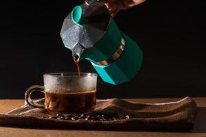 häll varmt svart kaffe från grön moka-kanna till klar kaffekopp med rök och kaffebönor på brun duk och träbord. fördelen med kaffekoncept. foto