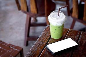 isgrönt te eller matcha latte med mjölkskum i en plastmugg med brunt sugrör och svart mobil eller smartphone och vit scen på ett träbord i kaféet. hälsosam dryck och dryck koncept. foto