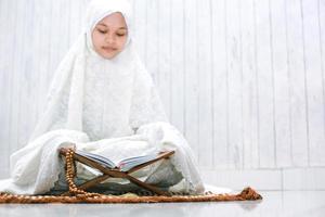 religiös ung asiatisk muslimsk kvinna som läser den heliga boken al-Koranen på bönemattan foto