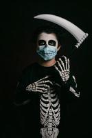 glad halloween, unge med medicinsk mask i en skelettdräkt med en lie foto