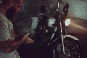 stilig brutal man med skägg står i sitt garage mot bakgrund av en motorcykel och tittar bort foto