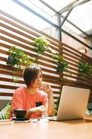 porträtt av självsäker mogen professionell kvinna i glasögon, en korall t-shirt som sitter på sommarterrass i café, använder bärbar dator för jobbet, skrattar glatt inomhus foto