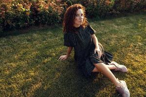 bedårande lockigt rödhårig tjej med fräknar i en klänning njuter av bra väder sittande på gräsmattan foto