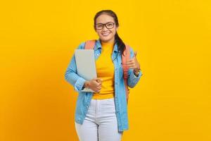 porträtt av glad ung asiatisk kvinna student i fritidskläder med ryggsäck som håller laptop och visar tummen upp med fingret isolerad på gul bakgrund. utbildning i högskolekoncept foto