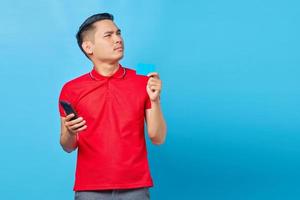 porträtt av eftertänksam ung asiatisk man som håller mobiltelefon och kreditkort isolerad på blå bakgrund foto