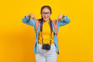 porträtt av glad ung resenär asiatisk kvinna med ryggsäck och kamera i jeanskläder medan du pekar på kopia utrymme med fingrar isolerad på gul bakgrund. flygresa koncept foto