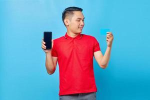 porträtt av glad ung asiatisk man visar tom skärm mobiltelefon och kreditkort isolerad på blå bakgrund foto