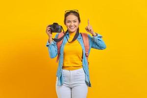 porträtt av leende ung asiatisk kvinna i jeanskläder med ryggsäck som håller professionell kamera och pekar uppåt med fingret isolerat på gul bakgrund foto