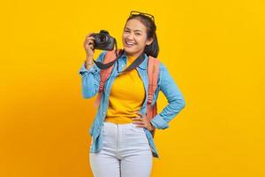 porträtt av glad ung asiatisk kvinna i jeanskläder med ryggsäck och visar professionell kamera isolerad på gul bakgrund foto