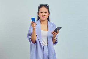 porträtt av förvirrad ung asiatisk kvinna som håller banktoken och håller smartphone över vit bakgrund. koncept pengaöverföring via internet foto