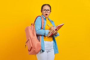 chockad ung asiatisk kvinna student i jeanskläder med ryggsäck, skriva anteckningar i anteckningsbok isolerad på gul bakgrund. utbildning i gymnasiekonceptet foto