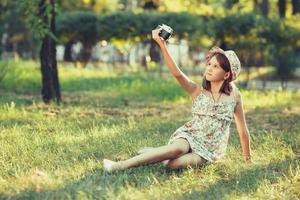 liten flicka spelas av fotokamera sitter på gräs i parken. göra selfie och fotografera världen runt foto