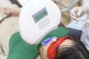 en kvinna som bär skyddsglasögon undersöker av stomatolog, tandblekning med ultraviolett lampa foto