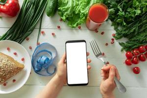 kvinnlig hand håller en smartphone på en bakgrund av ett bord med dietgrönsaker. plats för din text. foto