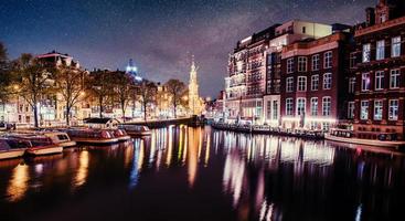 vacker natt i amsterdam. nattbelysning av byggnader och båtar nära vattnet i kanalen. foto