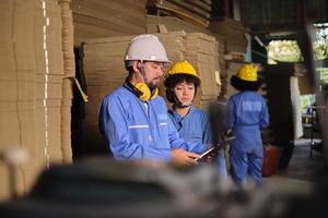 asiatisk manlig ingenjör i säkerhetsuniform och hjälm och kvinnlig arbetskollega inspekterar lager, lagerbeställning på fabrikslager, högar med stapling av kartongtillverkning, industriprodukthantering. foto