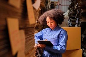 afroamerikansk kvinnlig leveransarbetare i säkerhetsuniform som använder en surfplatta för att kontrollera leveransordrar på paketlager, papperstillverkningsfabrik för förpackningsindustrin, logistisk transporttjänst. foto