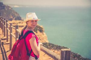 ung kvinna resenär med vit hatt och röd klänning som vistas nära trästaket vid klippan av cabo da roca cape roca foto