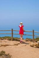 ung kvinna resenär med vit hatt och röd klänning som vistas nära trästaket vid klippan av cabo da roca cape roca foto