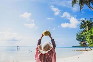 kvinna i sommarhatt håller kokosnöt på stranden med vacker blå himmel och havsbakgrund med kopia utrymme foto