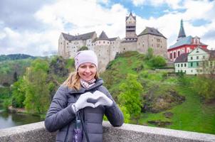 ung kvinna turist med grå jacka poserar, le och visa hjärta med fingrarna nära loket slott foto