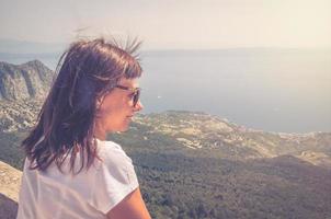 ung kaukasisk kvinna resenär med vit t-shirt och solglasögon tittar bort avstånd på Adriatiska havet foto