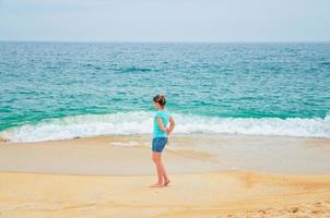 ung kvinna resenär gå ner sandstrand kusten och tittar på turkost vatten foto