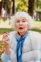 ledsen fashionabla äldre kvinna med grått hår i pälsrock utomhus röka cigarett. ohälsosam livsstil, ålder, gammalhet, missbruk, dålig vana koncept. foto