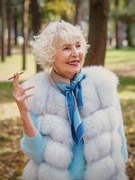 leende senior elegant snygg fashionabla kvinna med grått hår i päls utomhus röka cigarett. ohälsosam livsstil, ålder, positiva vibbar, gammalhet, missbruk, dålig vana koncept. foto