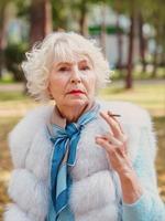 ledsen fashionabla äldre kvinna med grått hår i pälsrock utomhus röka cigarett. ohälsosam livsstil, ålder, gammalhet, missbruk, dålig vana koncept. foto