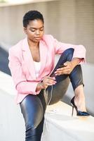svart affärskvinna som sitter utomhus med smartphone med hörlurar foto