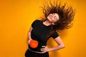 ung kvinna i fritidskläder med en orange mugg i handen är nöjd med kaffe hon har druckit och njuter av munterhet på morgonen och kastar håret bakåt. står isolerad på gul bakgrund foto