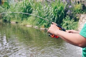 närbild av spinning med fiskerullen i handen, fiskekrok på linan med betet i vänster hand mot bakgrund av vattnet. foto