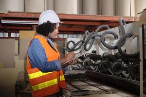 en afroamerikansk kvinnlig industriarbetare med hjälm kontrollerar maskinen och skriver inspekterade motordelar. professionell arbetar i en papperstillverkningsfabrik, underhåller produktionsutrustning. foto