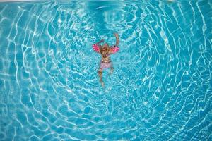 söt liten liten flicka simmar i turkos pool med uppblåsbara armar hjälper stöd, ovanifrån. foto