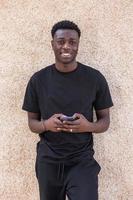 glad afrikansk amerikansk man meddelanden på smartphone nära betongvägg foto