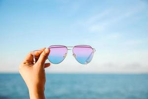 selektiv inriktning av hand som håller solglasögon med havet och blå himmel bakgrund foto