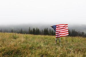 glad bedårande liten flicka ler och viftar med amerikanska flaggan. patriotisk helgdag. glad unge, söt liten barn flicka med amerikanska flaggan. usa firar den 4 juli. självständighetsdagen koncept. foto