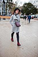 ung modell turist flicka i en grå rock och svart hatt med läder handväska på axlarna poserade på gatan i staden. foto