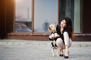 brunett zigenare tjej med yorkshire terrier hund poserade mot stora fönster hus. modell slitage på skinnjacka och t-shirt med ornament, byxor och skor med höga klackar. foto