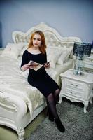 rödhårig tjej på svart klänning sitter på sängen och läser modetidning. fototonade instagramfilter. foto