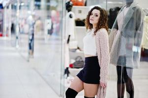 ung lockig modell tjej poserade på minikjol på stort köpcentrum nära skyltfönster med skyltdocka. foto