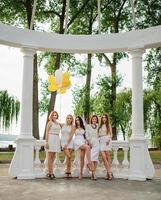 fem flickor med ballonger till hands bärs på vita klänningar på möhippor mot vita kolumner av båge. foto