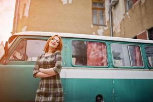 ung rödhårig flicka poserade på rutig klänning bakgrund gamla retro cyan minivan. foto