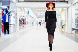 mode rödhårig flicka bär på svart klänning och röd hatt poserade på handel köpcentrum. fototonade instagramfilter. foto