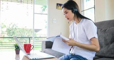 asiatisk glad kvinna student bära hörlurar studera online titta på webbseminarium podcast på laptop lyssna lärande utbildning kurs konferenssamtal och skriva anteckningar titta på laptop sitta på soffan hemma foto