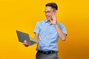 porträtt av glad ung asiatisk man asiatisk i glasögon med en bärbar dator, gör ett videosamtal, gestikulerande hej till vänner isolerad på gul bakgrund. affärsman och entreprenör koncept foto